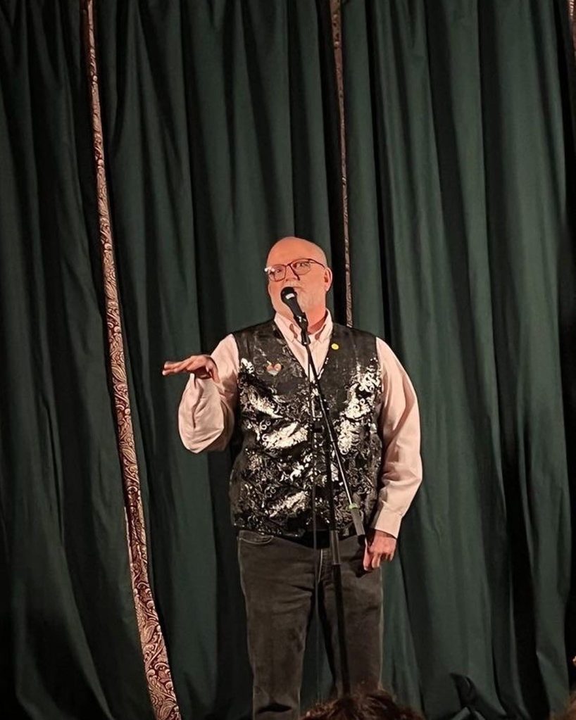 A picture of Mona Mae's first performance in Glasgow. Mona is presenting her gender as a male, but wearing a sparkly waistcoat, speaking into a microphone, and waving a limp wrist about. He/she looks VERY camp